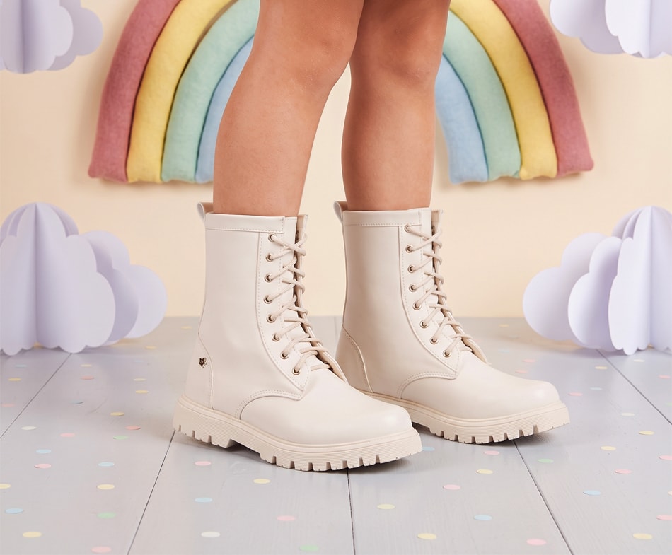 Pernas de criança calçando uma bota coturno infantil feminina na cor off-white (creme). O calçado possui sola tratorada e cadarços na mesma tonalidade.