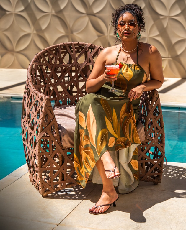 Mulher com óculos de sol e vestido verde com estampado de folhas, sentada numa cadeira de vime junto à piscina. Segura um cocktail e calça umas sandálias de salto baixo castanhas.