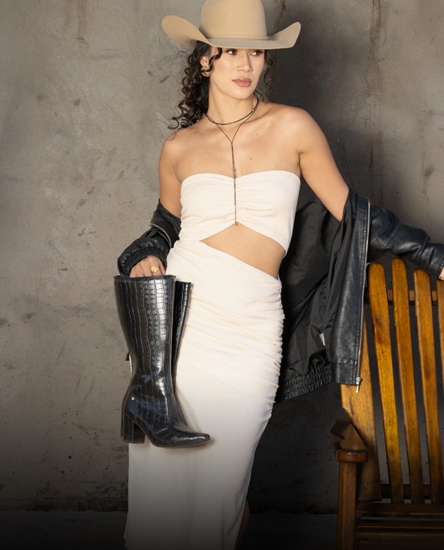 Mulher com chapéu e vestido bege segurando uma bota feminina preta de cano longo com textura croco e salto bloco.