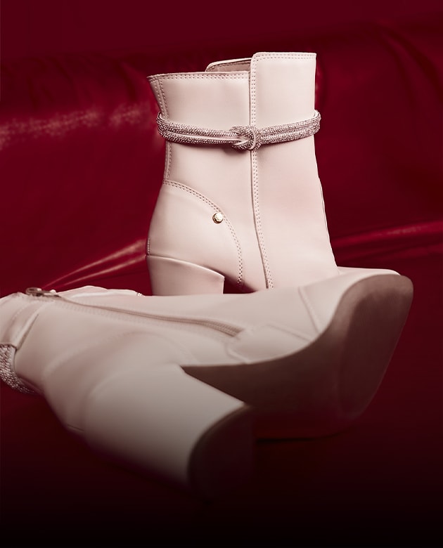 Bota feminina cano curto de salto bloco médio na cor off-white, destacando o detalhe luxuoso de tira cravejada de cristais strass com nó no tornozelo. O par está posicionado de forma conceitual sobre um fundo vermelho cereja acetinado com sombras dramáticas da campanha de inverno Domidona.
