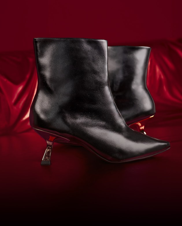 Par de botas femininas de cano curto pretas em couro macio com bico fino e salto taça kitten heel metalizado dourado. O calçado ganha destaque em um cenário luxuoso com drapeado de tecido vermelho brilhante e iluminação marcada de alta costura da campanha Domidona.