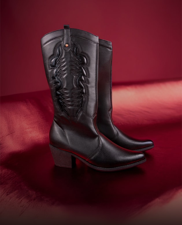 Bota feminina modelo texana western em couro preto com bordados pespontados detalhados no cano longo e bico fino. O calçado está posicionado em plano vertical sobre uma superfície acetinada vermelho escuro com iluminação dramática para o editorial de Dia dos Namorados Domidona.