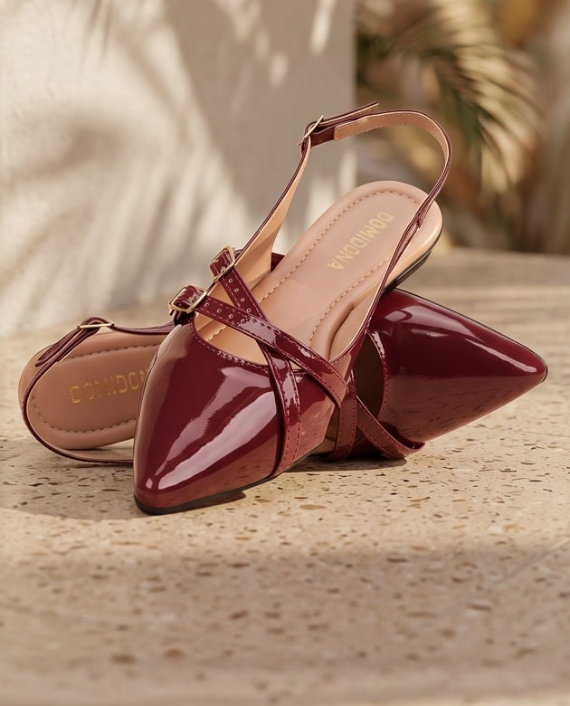 Par de sapatilhas slingback de bico fino na cor marsala em verniz, detalhadas com tiras cruzadas sobre uma superfície rústica.