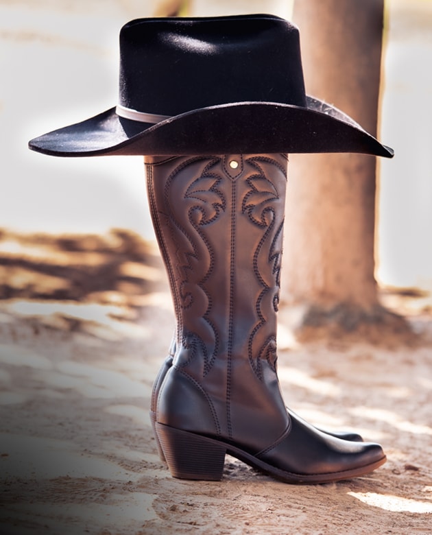 Bota texana feminina em tom escuro com bordados western tradicionais no cano. A bota está apoiada no chão de terra com um chapéu country preto descansando sobre ela