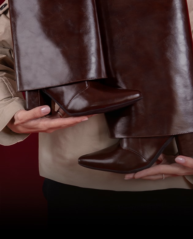 Bota feminina de cano alto com dobra sobreposta estilo slouchy cap na cor marrom chocolate, confeccionada em couro premium. Modelo posa segurando o par de botas vestindo casaco trench coat bege, sob fundo degradê vermelho escuro dramático para a campanha editorial de Dia dos Namorados Domidona.