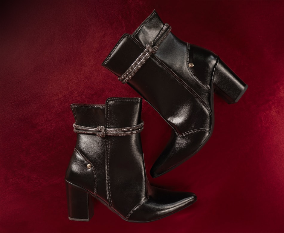 Vista diagonal superior de um par de botas femininas de cano curto pretas com bico fino e salto bloco médio, apresentando o detalhe sofisticado de tira de strass brilhante amarrada em nó no tornozelo. Fundo conceitual aveludado na cor vermelho vinho profundo com sombras suaves de inverno Domidona.