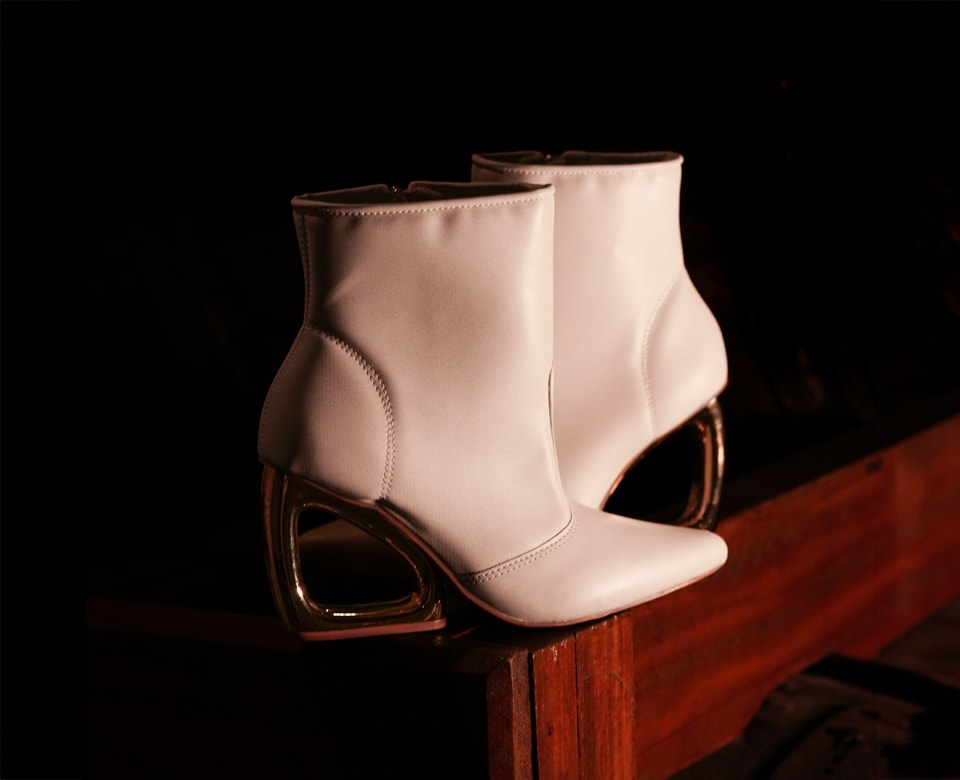 Bota feminina de cano curto e bico fino na cor off-white, destacando o exclusivo e elegante salto metálico polido de cor dourada com um recorte arquitetônico em forma da letra D vazado
