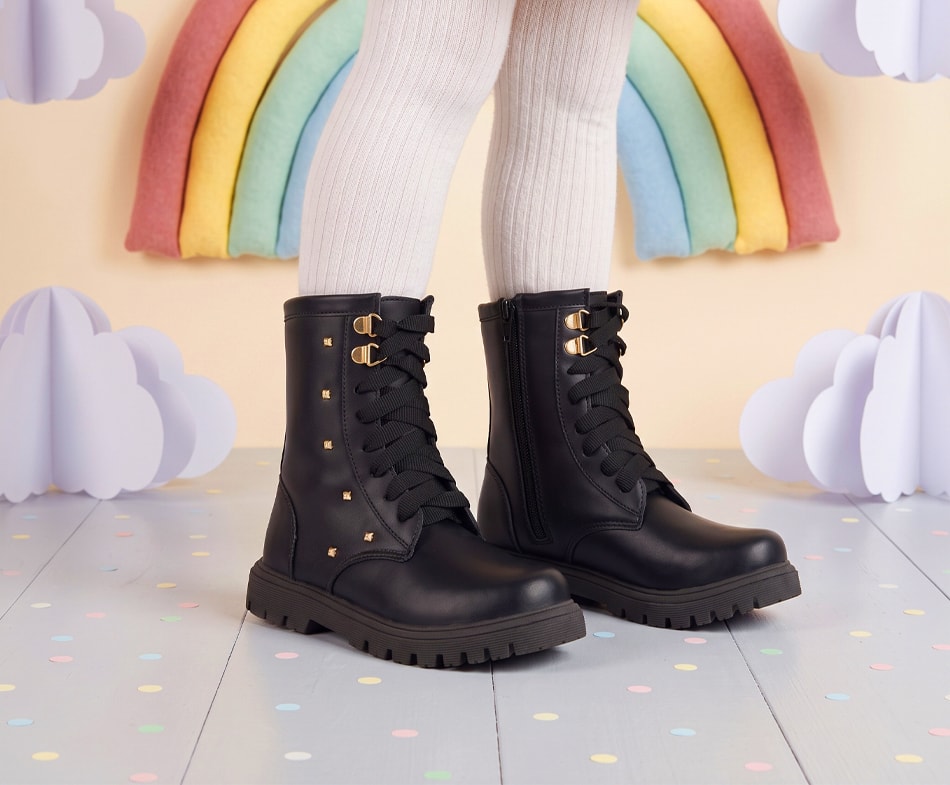 Menina vestindo meia-calça branca e calçando uma bota coturno infantil feminina preta. O modelo possui sola tratorada, cadarços largos e detalhes laterais em spikes dourados.