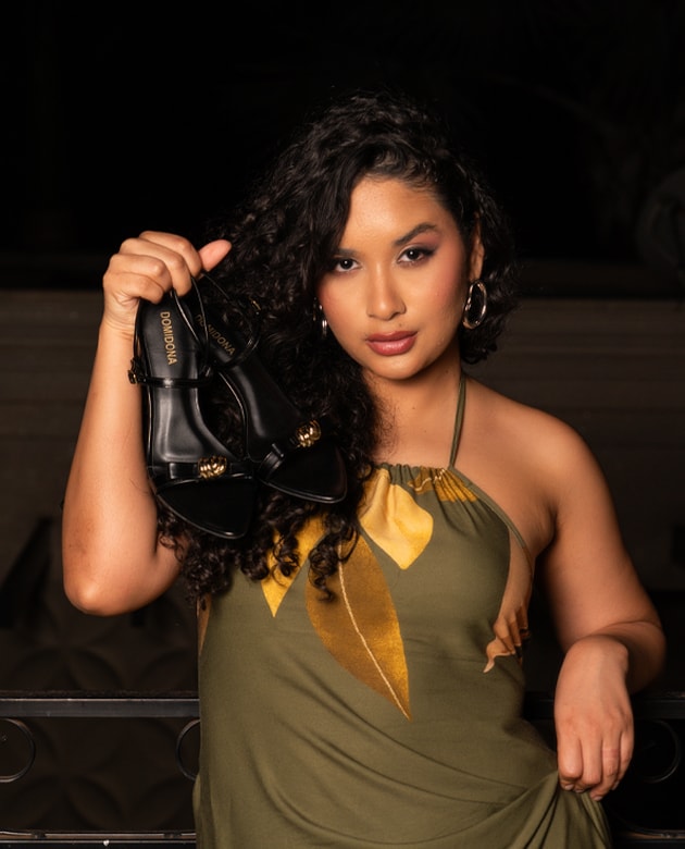 Mulher de cabelo encaracolado e vestido verde com estampado floral segura um par de sandálias pretas de salto com bico folha. As sandálias têm um pormenor dourado na tira frontal e a marca Domidona visível na palmilha. Fundo escuro.