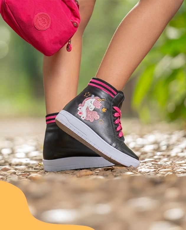 Detalhe de tênis cano alto infantil feminino na cor preta com estampa de unicórnio rosa brilhante e cadarço rosa. Modelo caminha em uma trilha de pedras