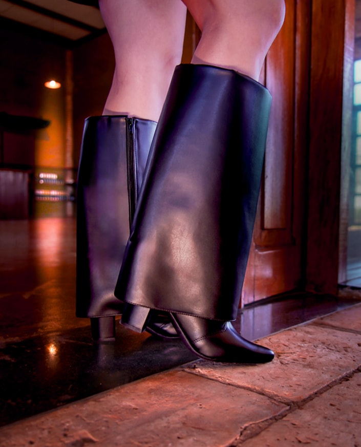 Foto de close-up focado nas pernas que vestem a Bota Capa Preta Domidona de couro polido. A imagem detalhada destaca o design exclusivo com cano alto estruturado e bico fino, sobre um piso de tijolos e com iluminação atmosférica quente de inverno no fundo