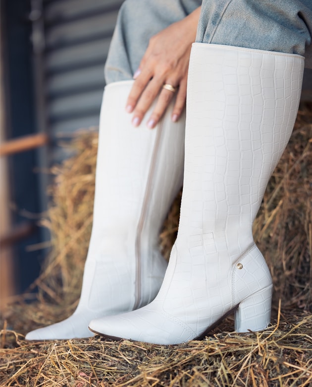 Bota feminina de cano longo branca com textura croco, bico fino e salto em bloco geométrico, apoiada sobre fardos de feno