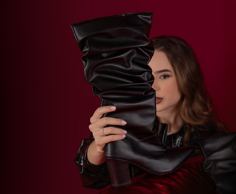 Modelo posa em enquadramento fechado segurando uma bota feminina slouchy de cano alto na cor preta, evidenciando as dobras do couro, bico fino e salto bloco elegante. Campanha editorial de moda com iluminação dramática e sombras fortes sobre fundo vinho para o Dia dos Namorados Domidona.