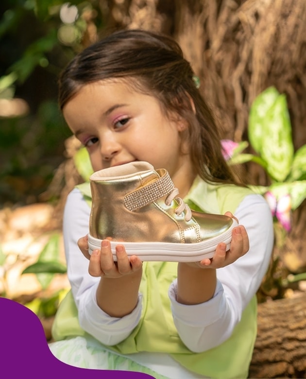 Menina sorrindo e segurando em destaque um tênis botinha infantil dourado metalizado com tira de strass brilhante no cano alto Menina Fashion