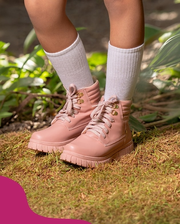 Criança usando bota estilo coturno infantil feminino na cor rosa com solado tratorado e meias brancas altas, pisando na grama. Coleção Menina Fashion