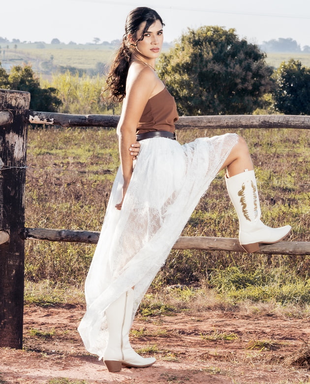 Bota western feminina de cano longo branca com recortes e bordados dourados no cano. Modelo em pé apoiada em uma cerca de madeira com saia longa branca e top marrom