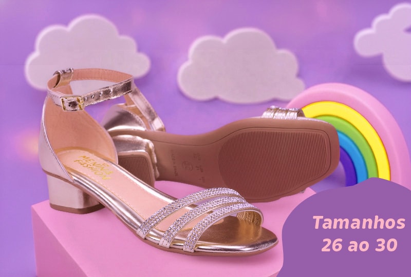 Sandália infantil feminina dourada metalizada de salto bloco baixo, com tiras frontais de strass e a palmilha com a logo "Menina Fashion". Apoiada sobre um bloco rosa com fundo lilás, nuvens e arco-íris. No canto inferior direito, uma etiqueta roxa destaca o texto "Tamanhos 26 ao 30".