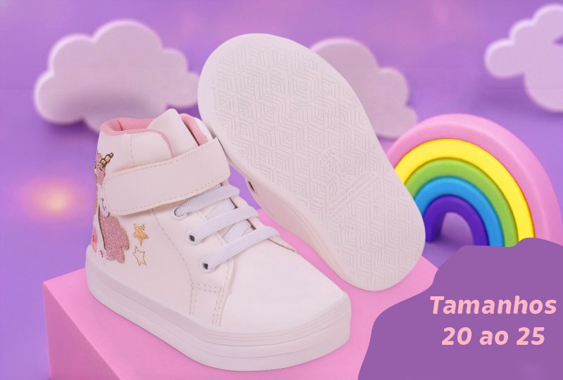 Par de tênis infantil feminino branco de cano alto com fecho de velcro e desenho de unicórnio com glitter. O calçado está apoiado sobre um bloco rosa com um cenário lúdico de fundo lilás, nuvens e arco-íris. No canto inferior direito, uma etiqueta roxa exibe o texto "Tamanhos 20 ao 25".